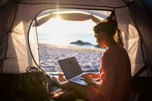 Side view of woman sitting in camping tent on the beach at sunrise, she using technology at sunrise.