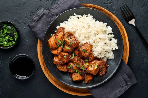 Photo of Spicy teriyaki chicken fillet pieces with rice, green onions and black sesame seeds on black plate on a dark slate, stone or concrete background. Top view with copy space.
