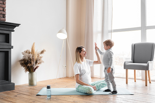 Young pregnant mother mom exercising on fitness mat with her little small kid son, giving high five. Sport, working out yoga with children. Future mother, pre-natal period, expecting a baby concept