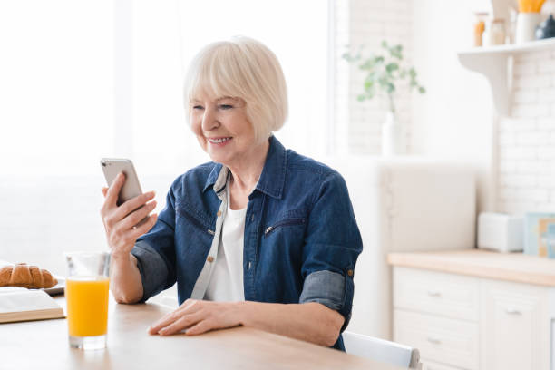 istock Happy old elderly senior aged mature woman using smart phone in kitchen having healthy breakfast. Smiling senior woman with orange juice checking messages, feed and social media on mobile phone. 1322712236