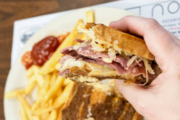 A patron hold a Rueben sandwich in one hand with the other half of sandwich and french fries blurred in the background. A patron hold a Rueben sandwich in one hand with the other half of sandwich and French fries blurred in the background. reuben sandwich stock pictures, royalty-free photos & images