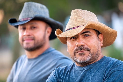Two hispanic mature agricultural male workers wearing cowboy hats looking at the camera smiling