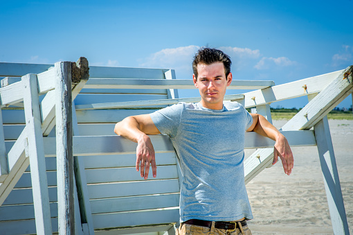 Man waiting for you. Wearing a gray t-shirt and arms resting on a wooden stick, a handsome young man is standing by a wooden structure on the beach, narrowing eyes, charmingly looking at you.