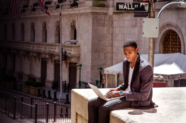 A black man is traveling, working in New York City A black man is traveling, working in New York City, sitting on a street by a vintage office building, reading, and working on a laptop computer. The Wall Street sign on the background. unidentifiable persons stock pictures, royalty-free photos & images