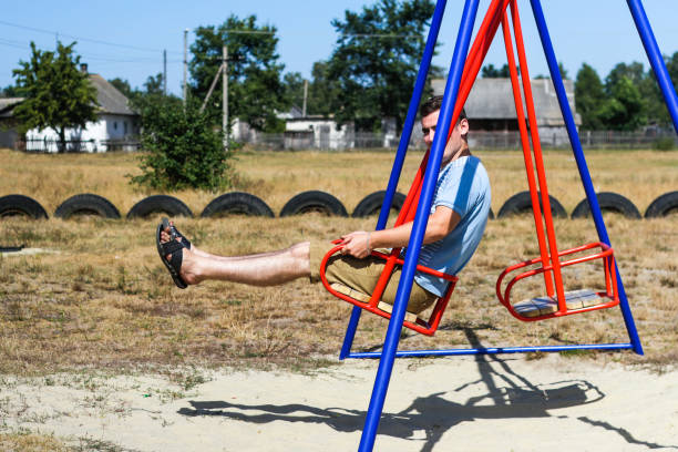 defocus young man swinging on swing on playground. countryside area. bright blue and red swing. kids summer game. guy raises legs and have fun on summer holiday. childlike adult. out of focus - freedom tire swing tire swing imagens e fotografias de stock