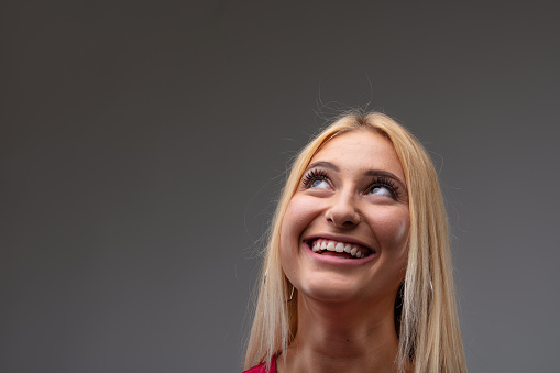 Cute young blond woman standing day dreaming with a happy smile and eyes raised to copyspace in a head shot portrait against a grey studio background