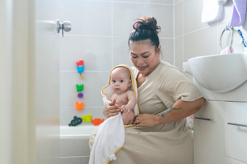 A beautiful mother dries her  baby daughter after bath time. The baby girl is wearing a hooded towel and looking at the camera. White bathroom setting. Tender moment between mother and daughter.