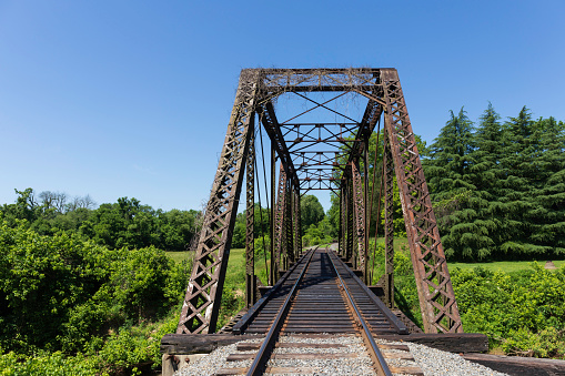Elkin, NC, USA-5 June 2021:  Image of a vintage steel railroad bridge, single point perspective, with tracks disappearing into lush green forest.  Horizontal image.