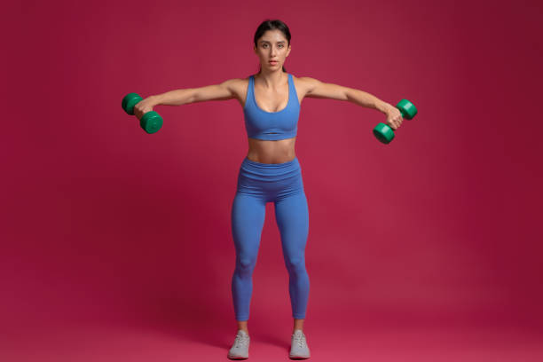 girl performing dumbbell lateral raise on maroon background - body building determination deltoid wellbeing imagens e fotografias de stock