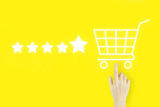 Photo of Customer service evaluation concept. Young woman's hand finger pointing with hologram Shopping cart and Five stars 5 rating on yellow background. Increase rating evaluation and classification concept.