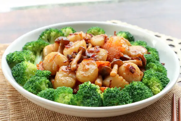 Photo of Stir fried Scallops with mushroom in  XO sauce of a white plate garnished with broccoli - close up view Chinese food on wood table