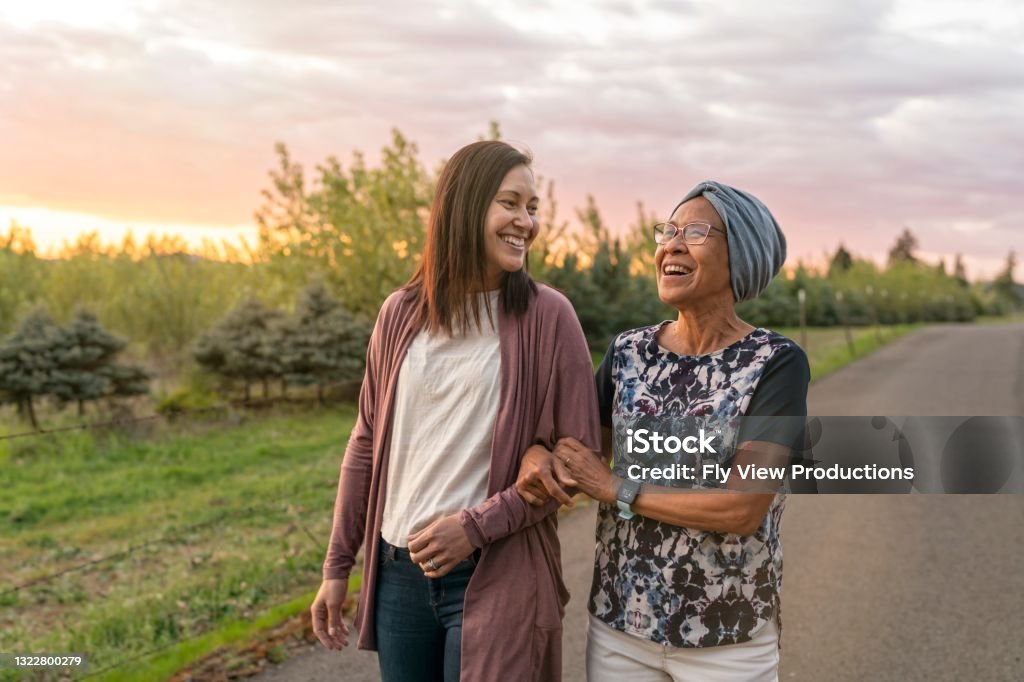 Beautiful mixed race mother and daughter relaxing outdoors together A senior woman with cancer walks with her adult daughter at sunset down a rural road. They are relaxing and staying active together. The affectionate pair are talking and walking with arms linked. Cancer - Illness Stock Photo