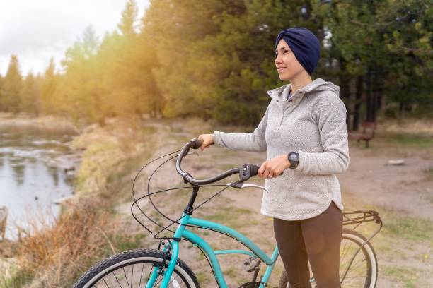 Portrait of young mixed race woman of Asian and Pacific Islander descent stopping along a river to soak in the beauty of nature while out on a relaxing bike ride. The smiling woman feels hopeful and confident after receiving news that cancer treatment was successful and she is now in remission.