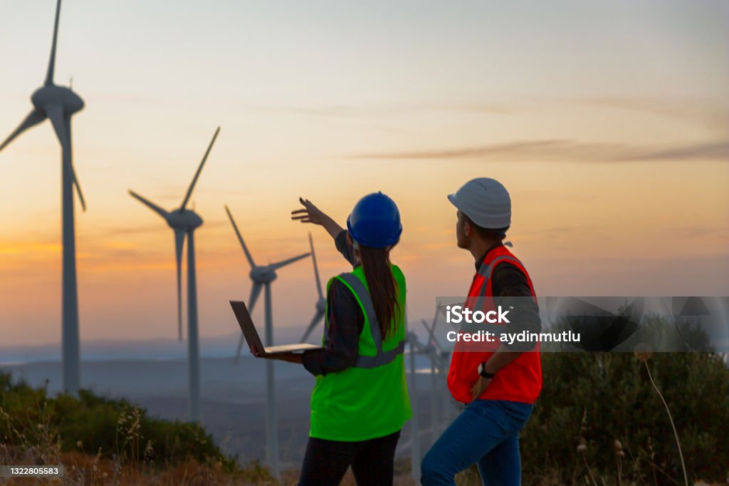 Young maintenance engineer team working in wind turbine farm at sunset Young electrical engineer woman and business man standing in front of wind turbines checking and working about technical problems and writes the results of measurements with laptop pc in wind power plant electric energy station. Wind Turbine Stock Photo