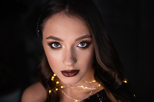 Close-up portrait of beautiful girl with an evening festive, professional make-up, bright, shiny garland around neck. Dark metallic background. Grunge texture of loft interior. Clean skin healthy face