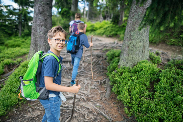 Mother and two sons are hiking in the forest. They are climbing up the trail on the tree roots. The boy is smiling back at the camera.\nPolish mountains Gorce.\nNikon D850