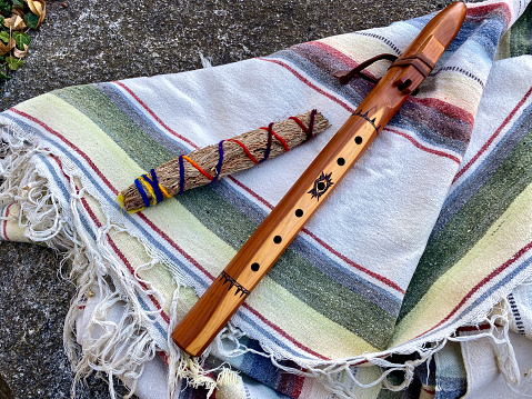Horizontal high angle closeup photo of a wooden handcrafted native American flute and a handmade sage smudge stick from the Taos Pueblo, lying on a woven, striped blanket from New Mexico