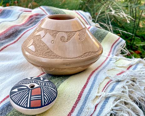 Horizontal closeup of two clay seed pots on a cotton striped fringed blanket from New Mexico. The large, handmade clay pot is etched with an animal shape. The small clay pot is painted with a pattern.