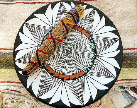 Horizontal high angle photo of a handmade smudge stick and a beautiful hand painted clay plate, on a striped, colorful cotton blanket, from New Mexico. The plate and the smudge stick are from the Taos Pueblo, Taos, New Mexico