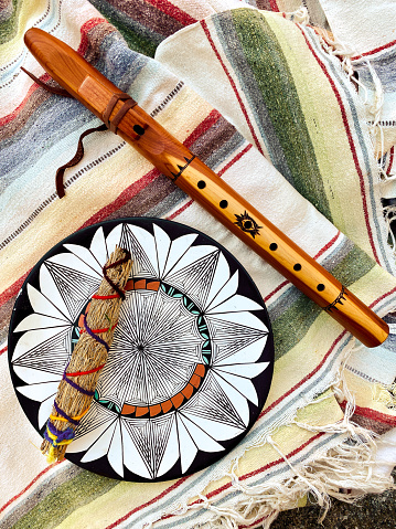 High angle closeup photo of a traditional native American wooden flute, a Taos Pueblo smudge stick handmade from local sage, a painted clay seed pot from Jemez Pueblo and a painted clay plate from Taos Pueblo lying on a colorful, striped, cotton blanket from New Mexico