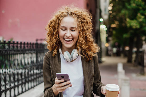 istock Young redhead woman is walking on the street and using mobile phone while holding reusable coffee cup 1322817975
