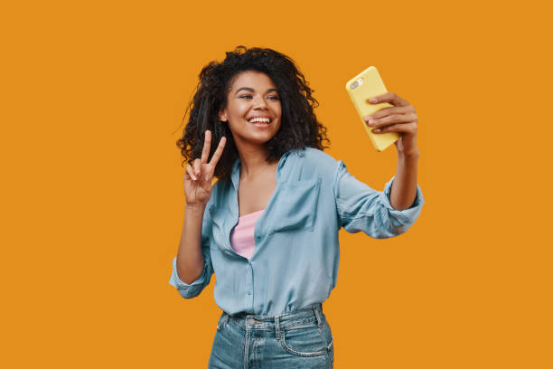 Beautiful young African woman in casual clothing taking selfie and smiling Beautiful young African woman in casual clothing taking selfie and smiling while standing against yellow background women taking selfies photos stock pictures, royalty-free photos & images