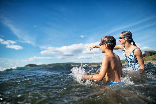 Brother and sister are having fun on the beach. Kids are walking in the shallow sea and looking at the sailboats.\nNikon D850