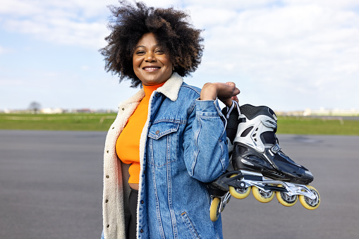 African woman carrying her roller skates outdoors on airtrip. Mature female skater carrying her inline skates on her shoulder and smiling.