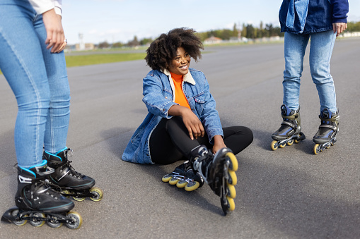 Tired looking african woman wearing roller skates sitting on road with her friends standing by. Woman getting tired after skating with friends on old runway.