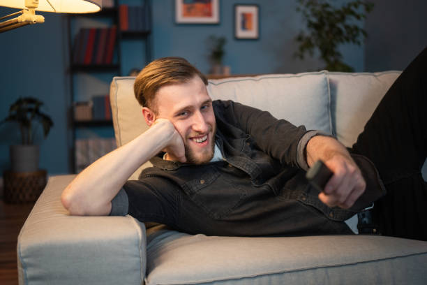 istock Smiling men is relaxing on sofa in living room, lying on couch lengthwise, supporting himself with arm under head, holding elbow on the armrest of sofa, holding remote control in hand to turn on TV 1322829966