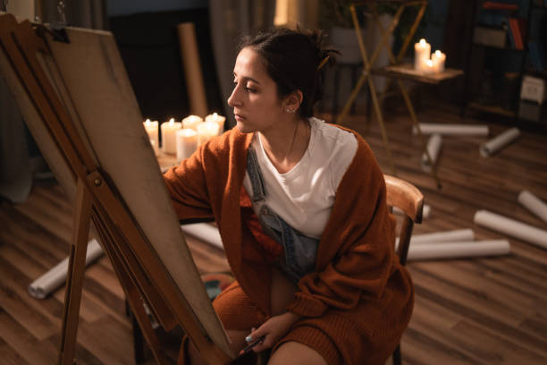 female sits in the middle of the room amidst scattered failed drawings and candles. the beautiful girl is focused on making the perfect painting, a pencil sketch - drawing sketch artist charcoal drawing imagens e fotografias de stock