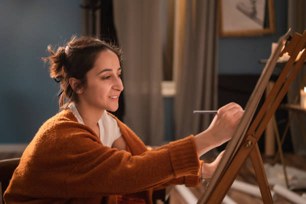 an art student sits in her home living room in the evening and draws a sketch of a tree, nature for a college credit. the passionate girl smiles, satisfied with painting - drawing sketch artist charcoal drawing imagens e fotografias de stock