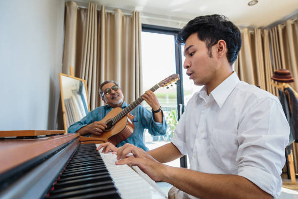 Asian adult son enjoy playing piano and senior father playing guitar together in living room at home Asian adult son enjoy playing piano and senior father playing guitar together in living room at home father and son guitar stock pictures, royalty-free photos & images