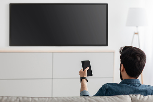 Young man watching television with blank screen and using smart TV remote control application on smartphone, back view. Select movie stream, change channel or settings in the menu