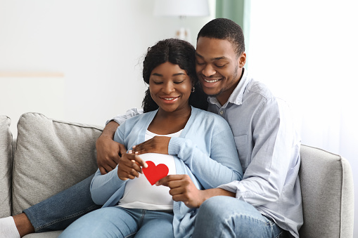 Cheerful expecting couple sitting on couch at home, holding red paper heart over pregnant lady big belly, young african american family waiting for baby, copy space. Parenthood, pregnancy concept