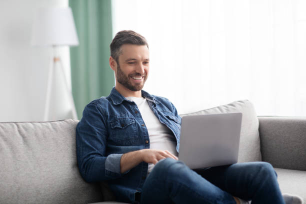 homme souriant utilisant un ordinateur portable, ayant un emploi à temps partiel à la maison - laptop sofa men computer photos et images de collection