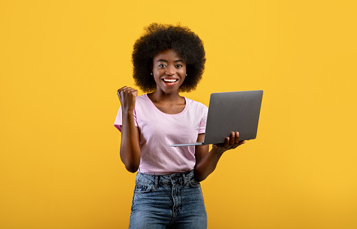 Euphoric black lady celebrating success with laptop, raising clenched fist over yellow background. Joyful woman holding computer and smiling to camera, cheering victory and success