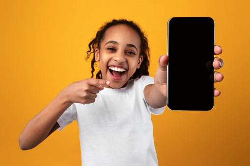 Excited Black Girl Pointing At Phone Blank Screen Showing Smartphone To Camera Recommending Application Over Yellow Studio Background. Great Mobile App For Cellphone Concept. Mockup