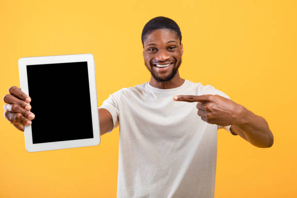 istock Positive african american guy pointing at tablet with blank black screen on yellow background, space for mockup 1322841165