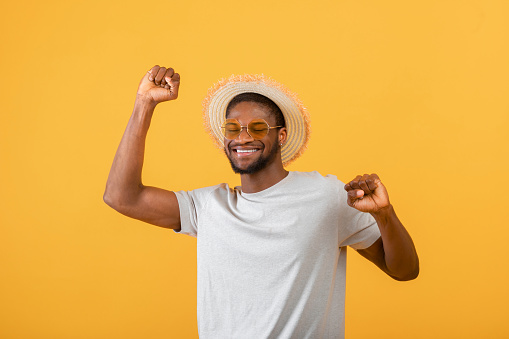Joyful african american guy in casual clothes, straw hat and sunglasses on yelow background, studio shot. Millennial black man ready for beach vacation, wearing summer outfit