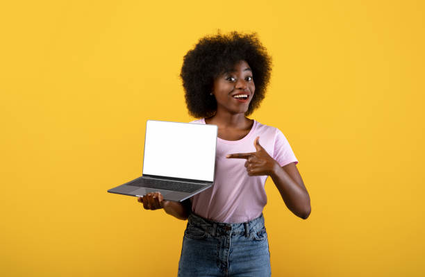 istock New website. Happy black lady holding laptop computer with blank screen for mockup and pointing on it, yellow background 1322842460