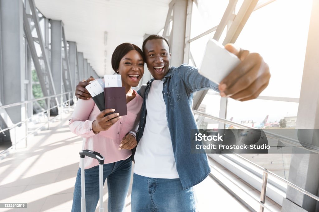 Ready For Adventures. Cheerful Black Couple Taking Selfie With Smartphone In Airport Ready For Adventures. Portrait Of Cheerful Young Black Couple Taking Selfie With Smartphone In Airport Terminal, Posing At Camera Together, Holding Passports And Tickets In Hands, Free Space Airport Stock Photo
