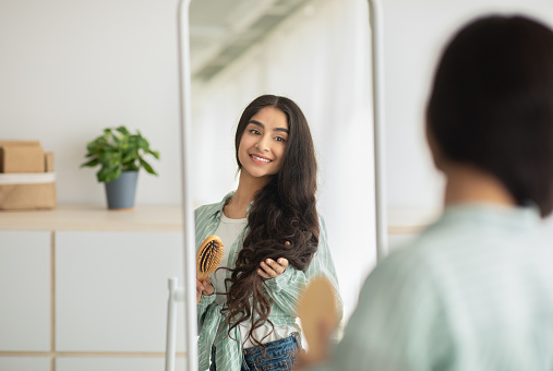 Cheerful Indian woman brushing her long beautiful hair with wooden brush in front of mirror at home. Pretty young lady taking care of her dark locks. Hairdressing and styling concept