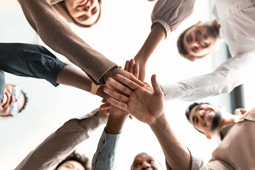 Teambuilding, Teamwork And Unity Concept. Below view of diverse group of smiling people putting their hands together, standing in circle. Multiethnic colleagues celebrating collaboration and alliance