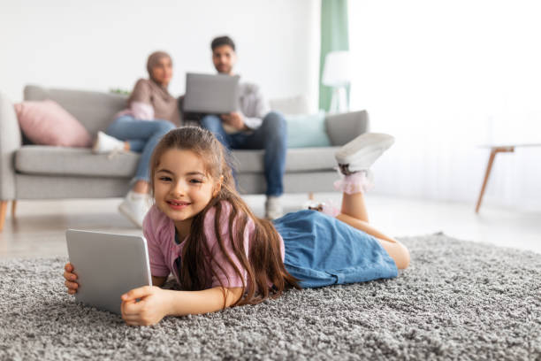 istock Pretty eastern girl using tablet, lying on floor carpet and smiling at camera, parents relaxing on sofa with laptop 1322843046
