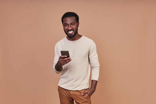 Handsome young African man in casual clothing using smart phone and smiling while standing against brown background