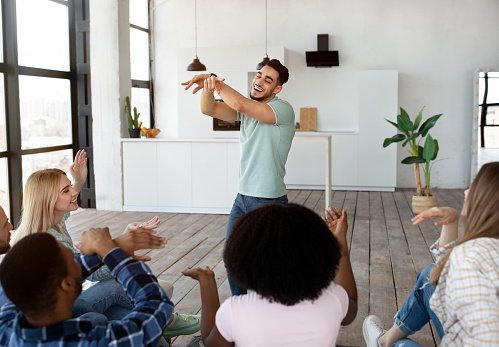 Group of happy multiracial friends playing charades guessing game at home. Cool millennial students showing pantomime t oeach other, having social gathering, enjoying each other's company