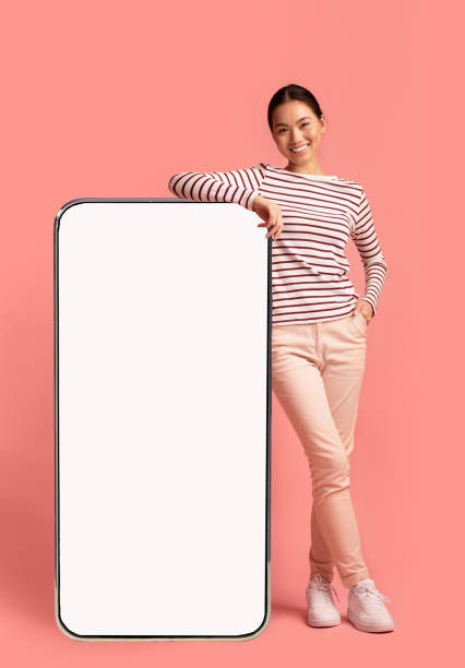 Mobile Offer. Smiling Asian Woman Leaning At Big Smartphone With White Screen Mobile Offer. Beautiful Smiling Asian Woman Leaning At Big Smartphone With Blank White Screen, Showing Free Copy Space For Your Design Or Advertisement, Posing Over Pink Background, Mockup Image leaning stock pictures, royalty-free photos & images