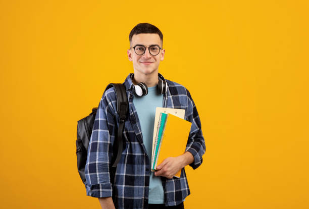 Education concept. Positive young man with backpack, headphones and notebooks posing on orange background
