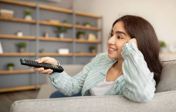 istock Lazy weekend. Lovely Indian lady with remote control watching TV while sitting on couch indoors, panorama 1322845711